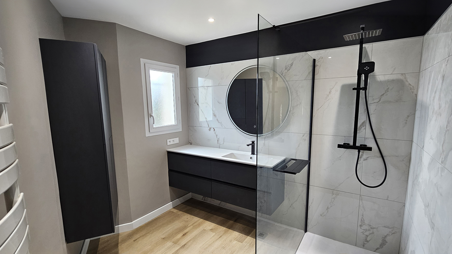 Salle de bain moderne rénovée avec douche italienne, meuble suspendu noir, grand miroir rond et robinetterie noire design, parquet clair et murs effet marbre blanc